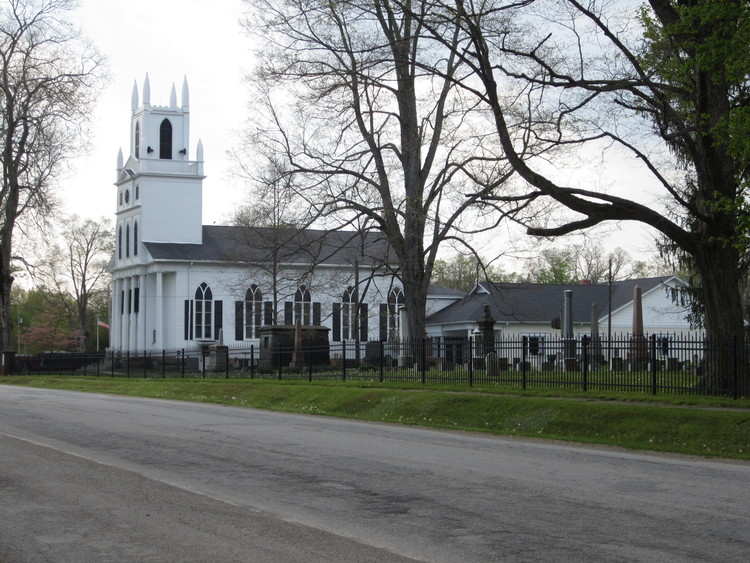 A historic white church with a tall steeple, surrounded by trees and a cemetery, is enclosed by a black iron fence.